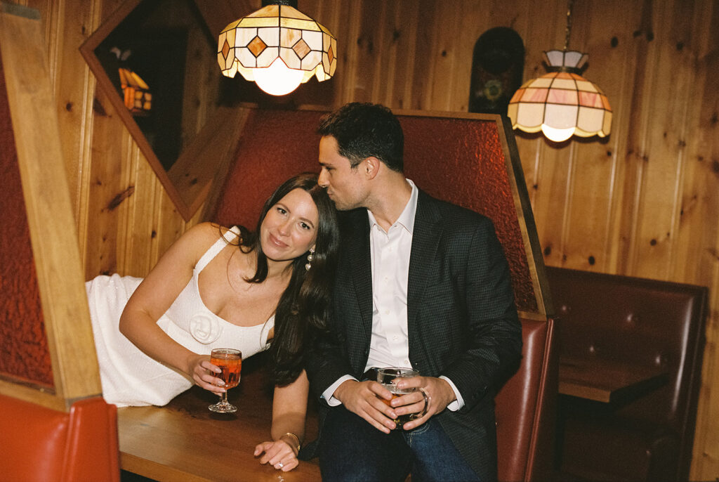 Candid engagement photo of a couple sharing drinks in a vintage wood-paneled Atlanta dive bar, captured in a nostalgic, film-style aesthetic by a documentary wedding photographer.