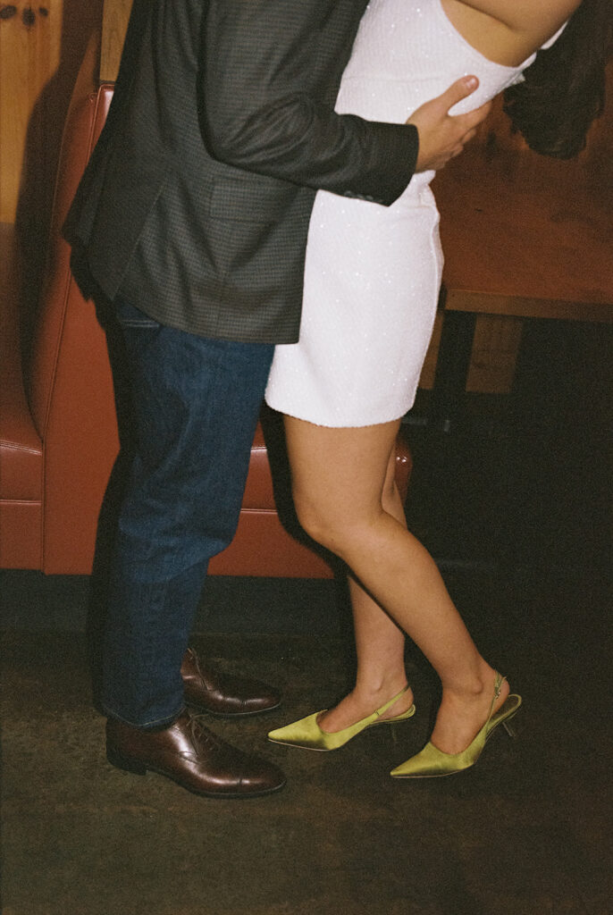 Candid engagement photo of a stylish couple in Atlanta, featuring the groom-to-be in a dark blazer and brown dress shoes and the bride-to-be in a sparkly white dress with green heels, captured in a vintage diner setting by a documentary-style wedding photographer.