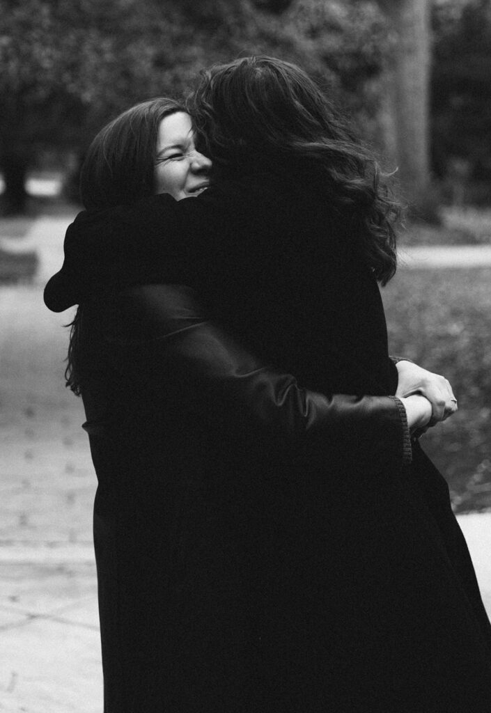 Two women wrapped in a joyful embrace, captured in a tender black and white moment. One beams with a scrunched-up smile as they hold each other close, love and laughter radiating between them—an honest, unscripted glimpse of queer joy and connection.