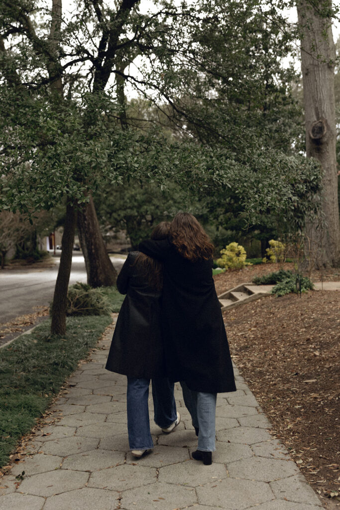 Two women walk hand-in-hand down a quiet residential street. They are both wearing long black coats and blue jeans, with one in heeled boots and the other in sneakers. The woman on the right looks back with a smile, while the other gestures playfully forward. Trees and houses frame the overcast scene.