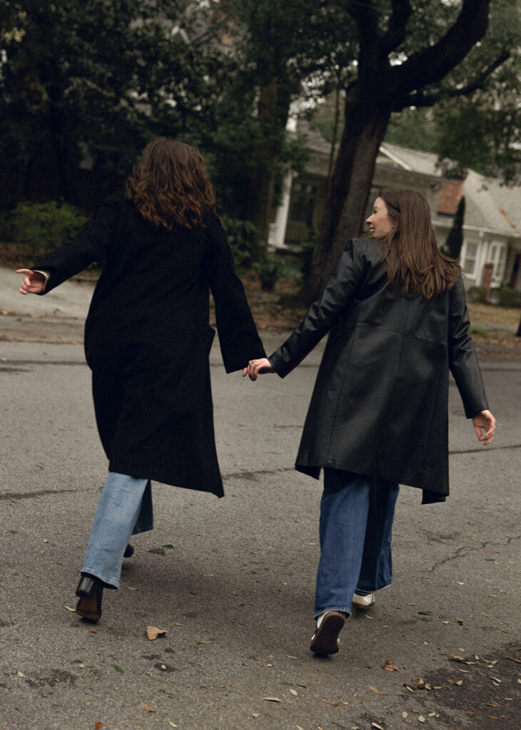 A joyful LGBTQ+ couple walks hand-in-hand down a residential path in Inman Park, Atlanta, surrounded by spring foliage. The image captures movement and intimacy in a documentary photography style.”