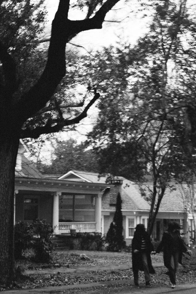 A black-and-white image captures the same couple walking away on a leaf-strewn sidewalk in a tree-lined neighborhood. Their figures are small in the frame, holding hands and mid-step, with large trees and vintage homes creating a dreamy, nostalgic atmosphere.
