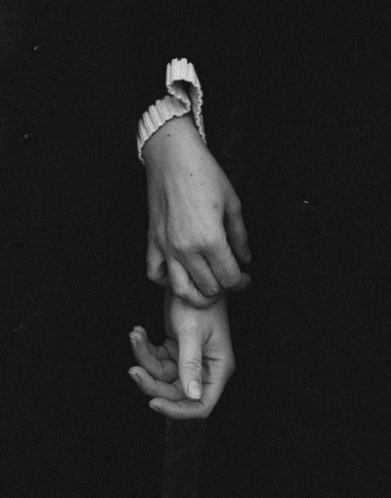 Close-up of two women’s hands resting together, each wearing a simple, meaningful ring. A quiet, intimate frame that speaks volumes—two fiancées grounding themselves in one another, wrapped in the stillness of queer love and commitment.