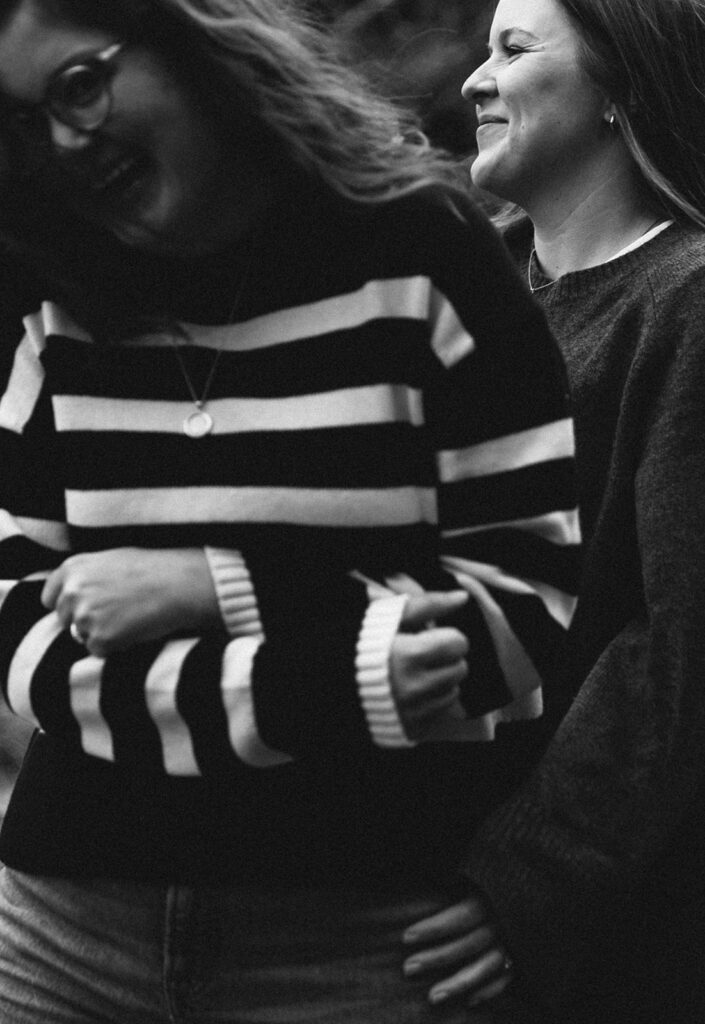 In a close-up black-and-white image, the couple laughs together. One woman wears a striped sweater and glasses, her face partially obscured by motion blur, while the other smiles with eyes closed, wearing a dark knit top. Their joyful expressions fill the frame.