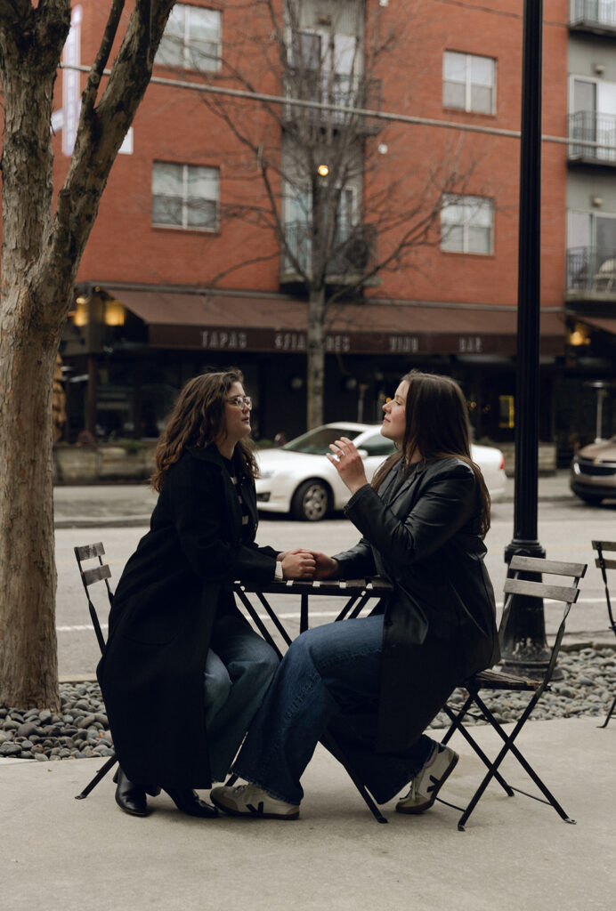 The couple sits at a small café table on a city sidewalk, holding hands and engaged in animated conversation. One woman wears a black coat and glasses, while the other wears a black leather jacket and sneakers. Behind them, a red brick building with a tapas bar sets an urban tone.