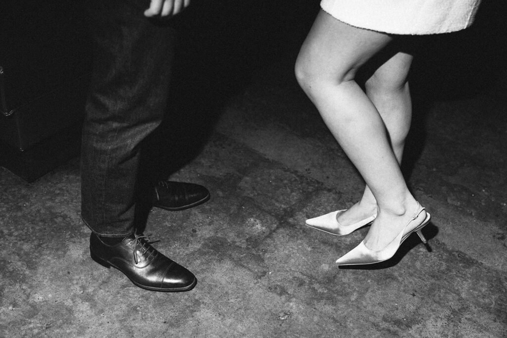 A candid black and white detail shot of the engaged couple’s feet—he in polished dress shoes, she in elegant heels—on a concrete floor.