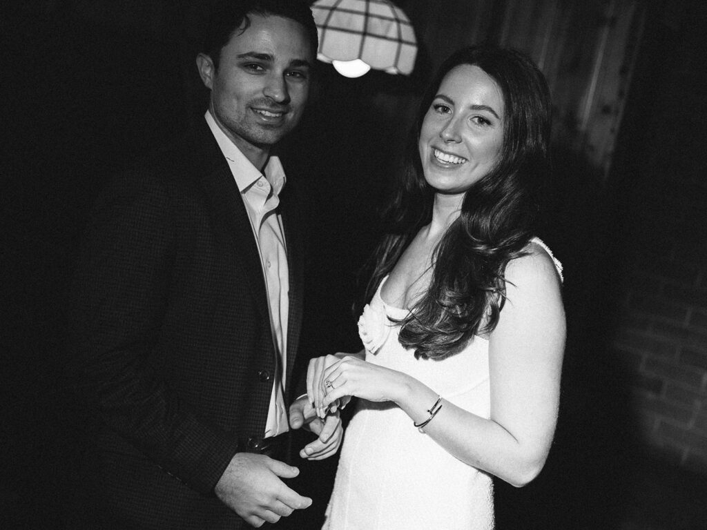 A joyful engaged couple smiling at the camera in a black and white portrait, taken during their documentary-style engagement shoot.