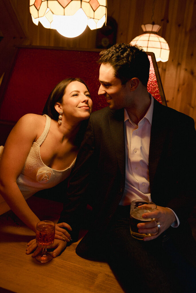Candid engagement photo of a couple sharing a romantic moment at a vintage diner in Atlanta, with the bride-to-be in a textured white dress and statement earrings leaning toward her fiancé, both illuminated by warm pendant lighting—captured by an Atlanta documentary wedding photographer.
