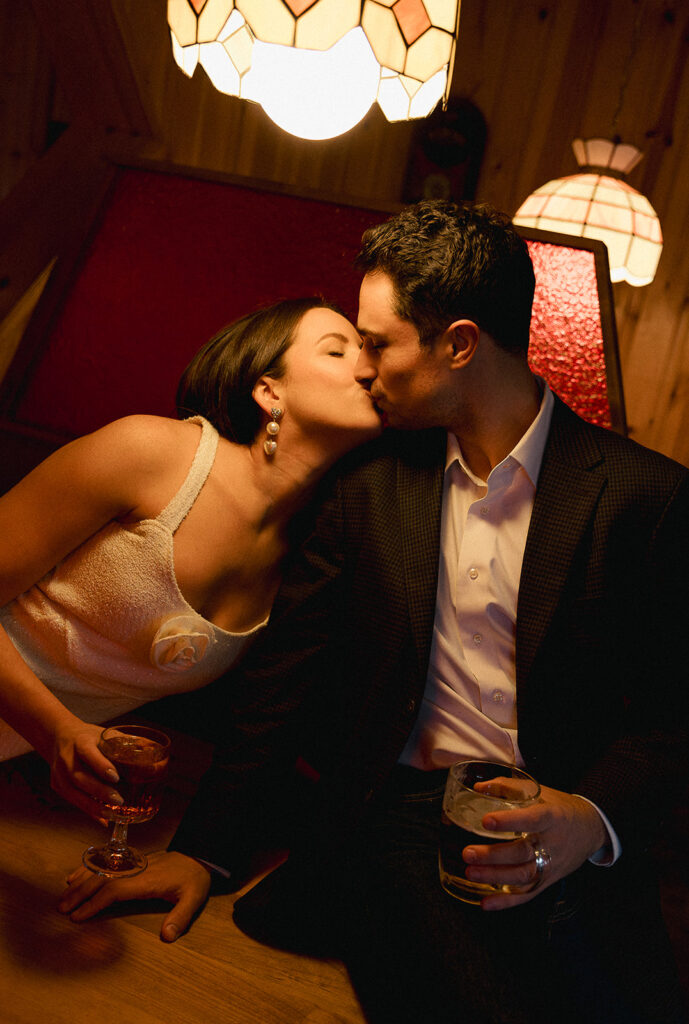 Documentary-style engagement photo of a couple sharing a candid kiss in a cozy restaurant booth under warm vintage pendant lights.