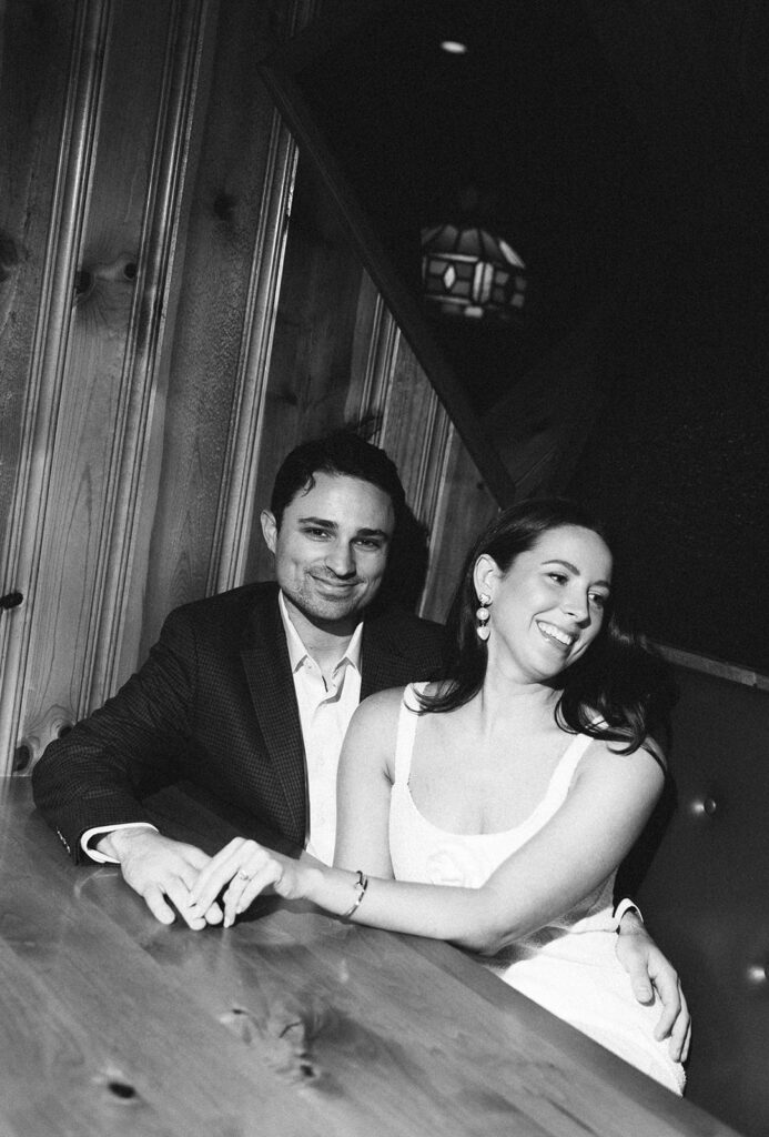 Black and white documentary engagement photo of a couple laughing and holding hands across a wooden booth table, capturing a candid moment in a vintage restaurant setting.
