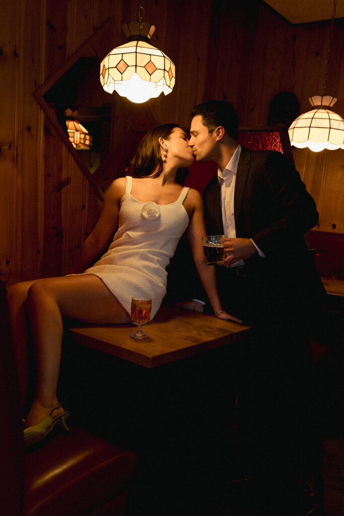 Documentary-style engagement photo of a couple sharing a candid kiss in a cozy restaurant booth under warm vintage pendant lights.