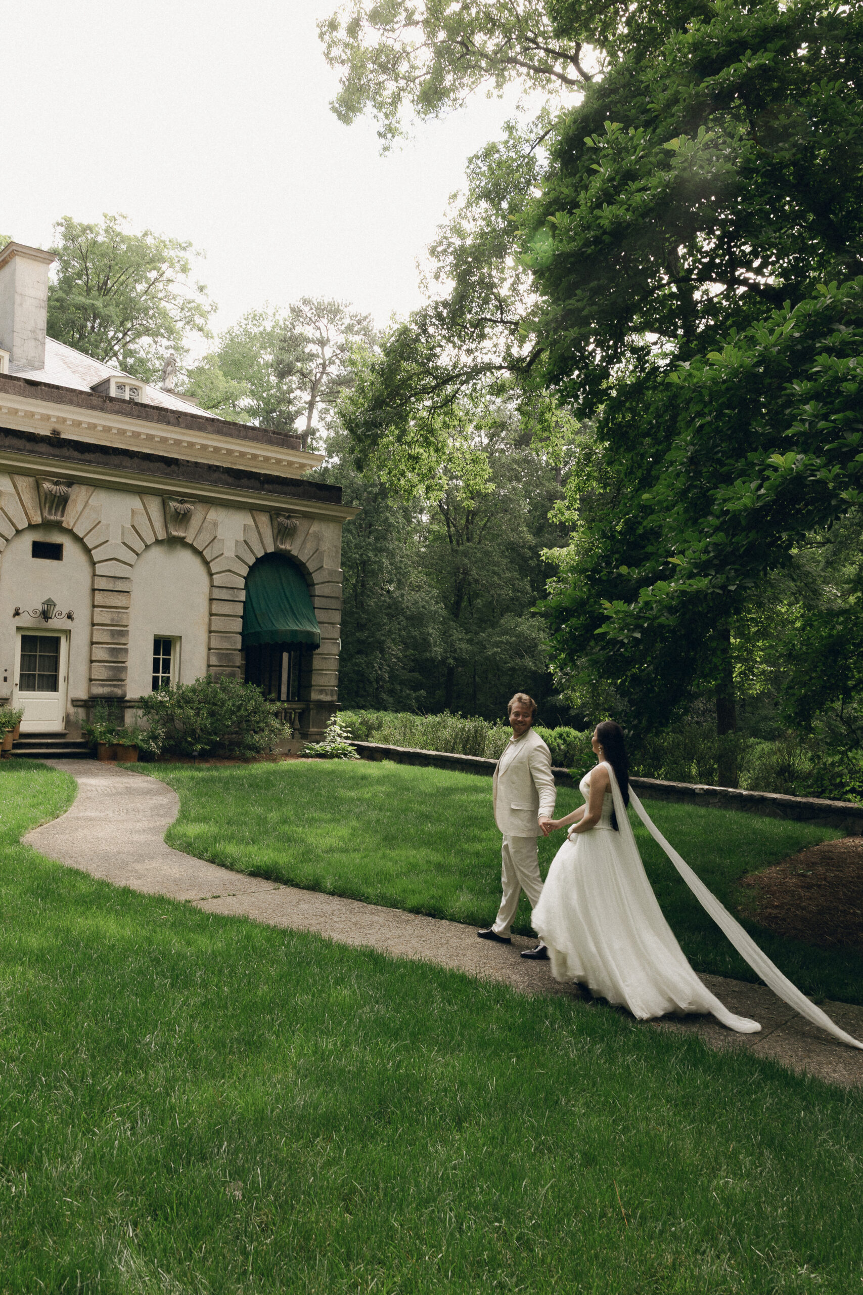 The couple walks hand in hand along a winding garden path beside the Swan House, the groom smiling back at the bride as her long veil trails behind her in the soft afternoon light.
