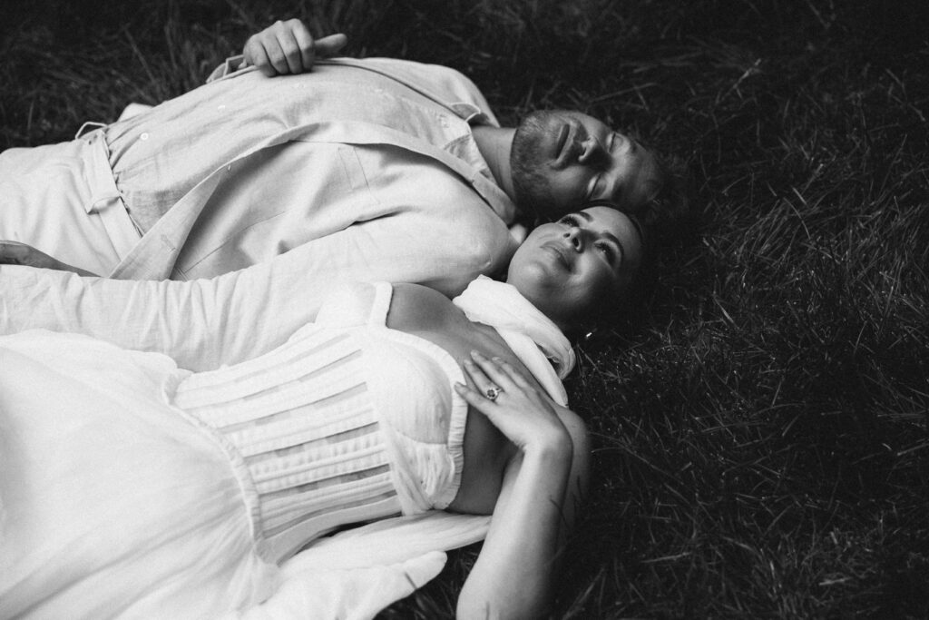 Black and white image of a bride and groom lying side by side on the grass, gazing peacefully upward, exuding quiet joy and connection.