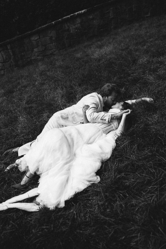 Black and white photo of a bride and groom lying on the grass, sharing an intimate kiss with a stone wall in the background.
