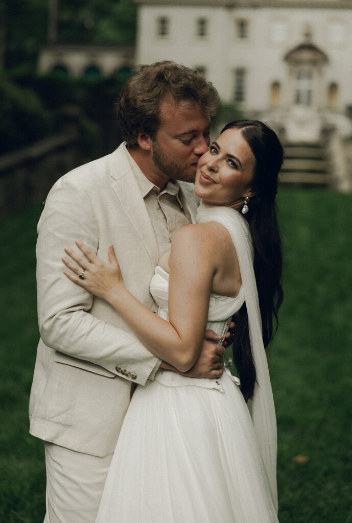 Bride and groom embracing on a lush lawn in front of an elegant estate; the groom kisses the bride's cheek as she smiles warmly at the camera.