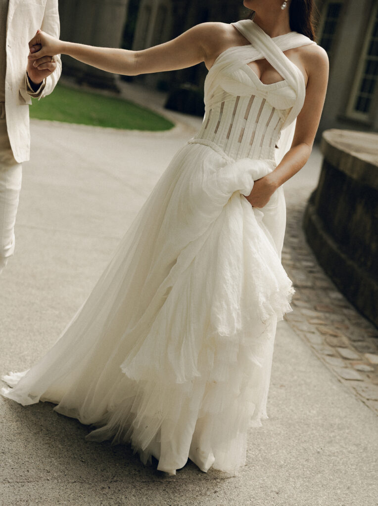 Close-up of a bride in a modern, sculptural wedding gown with a corset bodice and layered tulle skirt, holding hands with her groom as they walk outdoors.