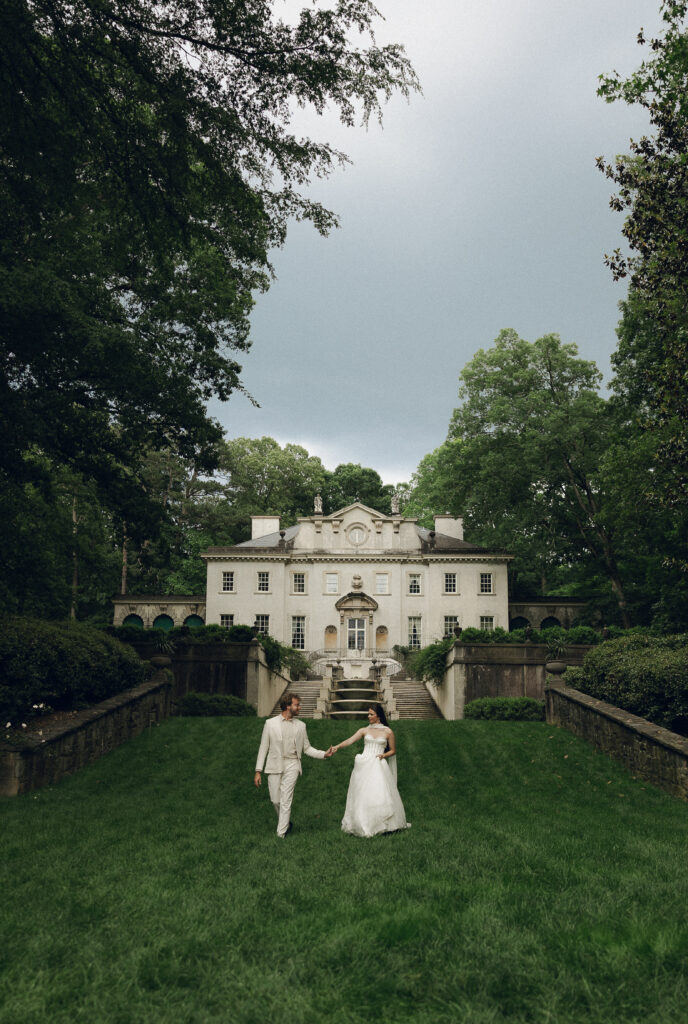 A couple walks hand in hand down the grand staircase of the Swan House, framed by lush greenery and classical architecture under a bright blue sky.