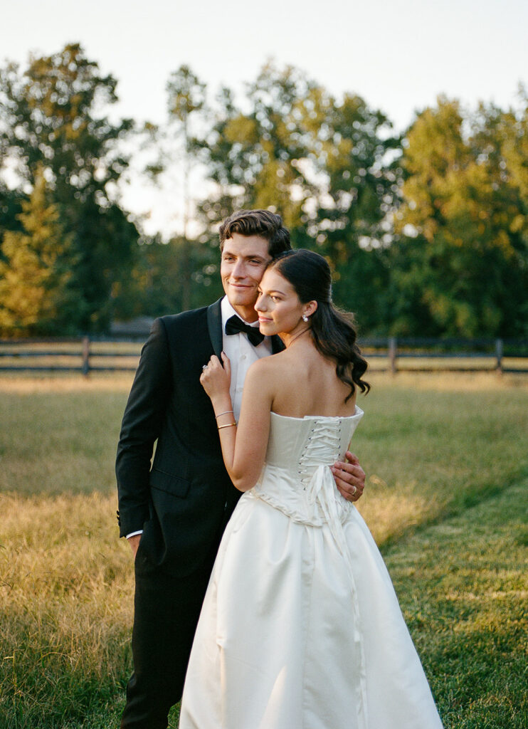 Bride and groom standing in a sunlit field at a private estate in Milton, Georgia. The groom wears a classic black tuxedo and the bride wears an elegant satin gown with a lace-up back, captured on film by wedding photographer Kate Phillips.