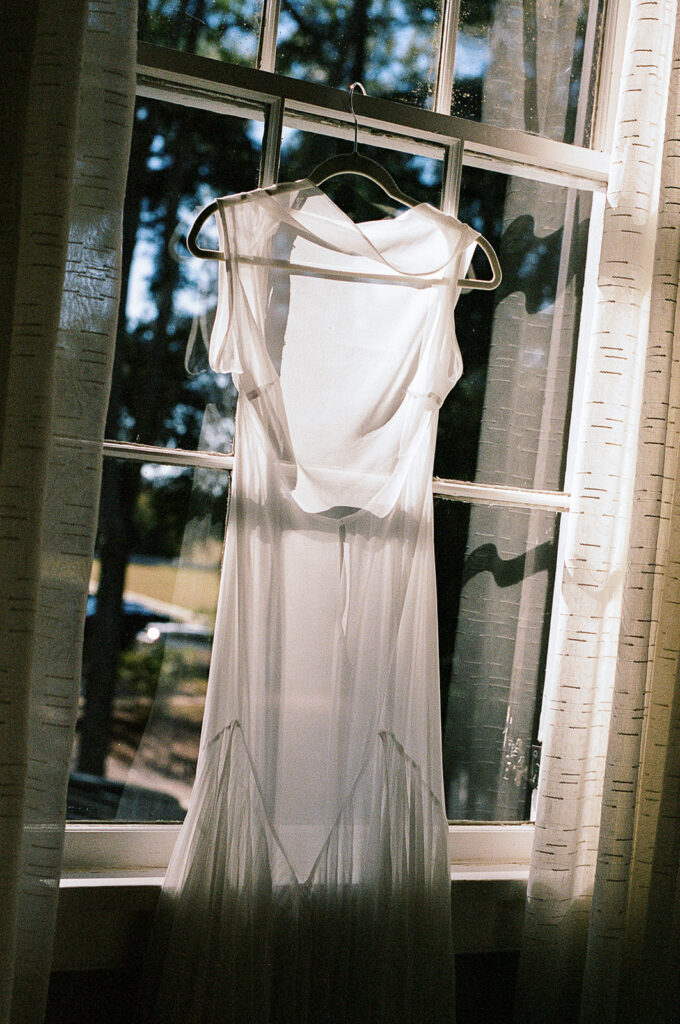 Wedding dress hanging in a sunlit window on film, captured during a quiet morning before the ceremony