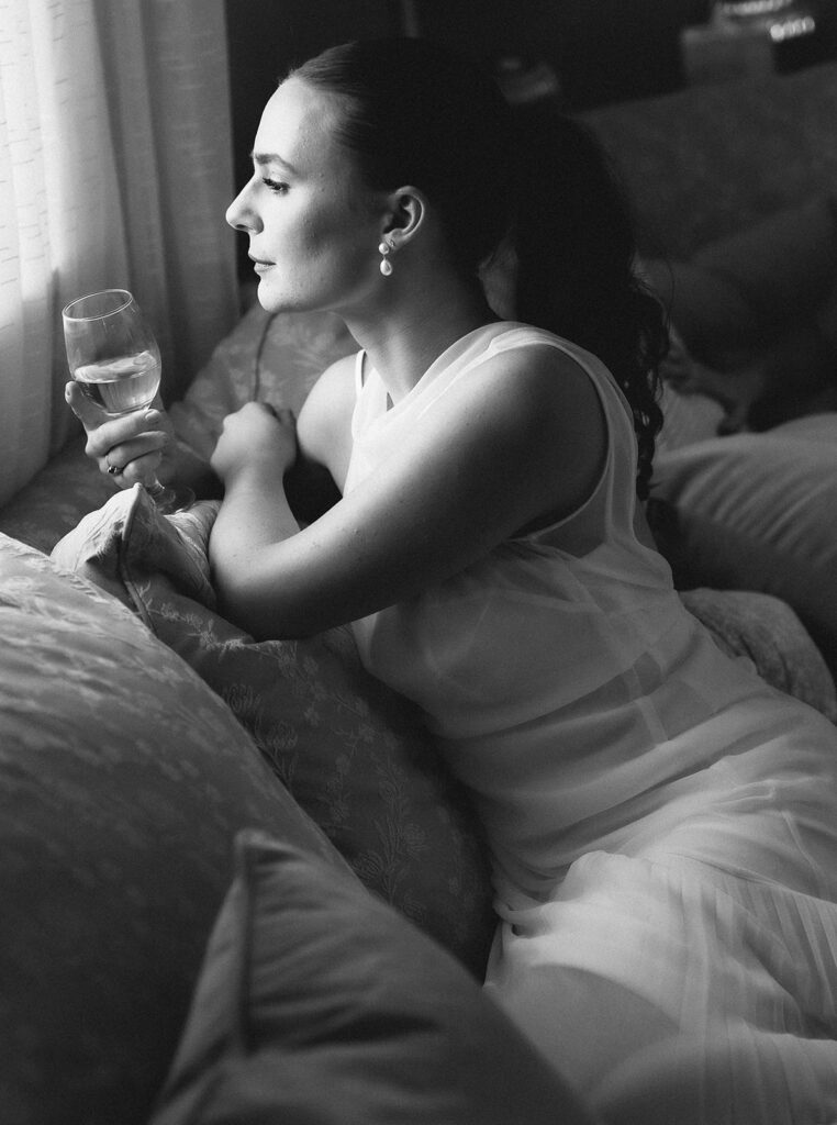 Black and white portrait of a bride holding a glass of wine while looking out the window during a peaceful pre-ceremony moment