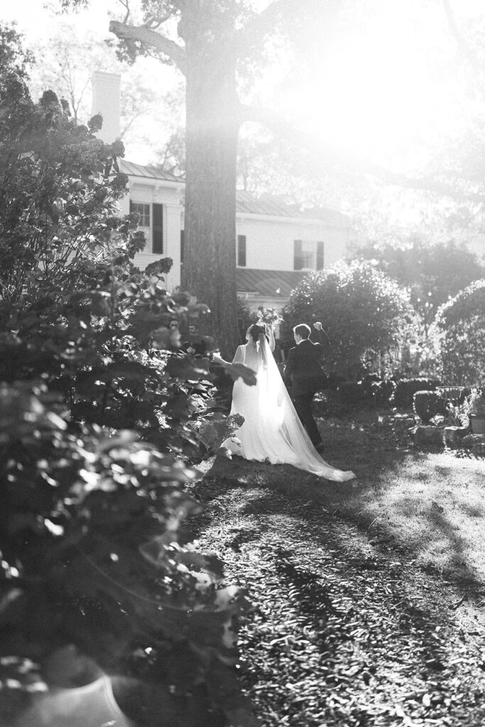 Black and white photo of a bride and groom walking through a sunlit garden with soft haze and natural foliage