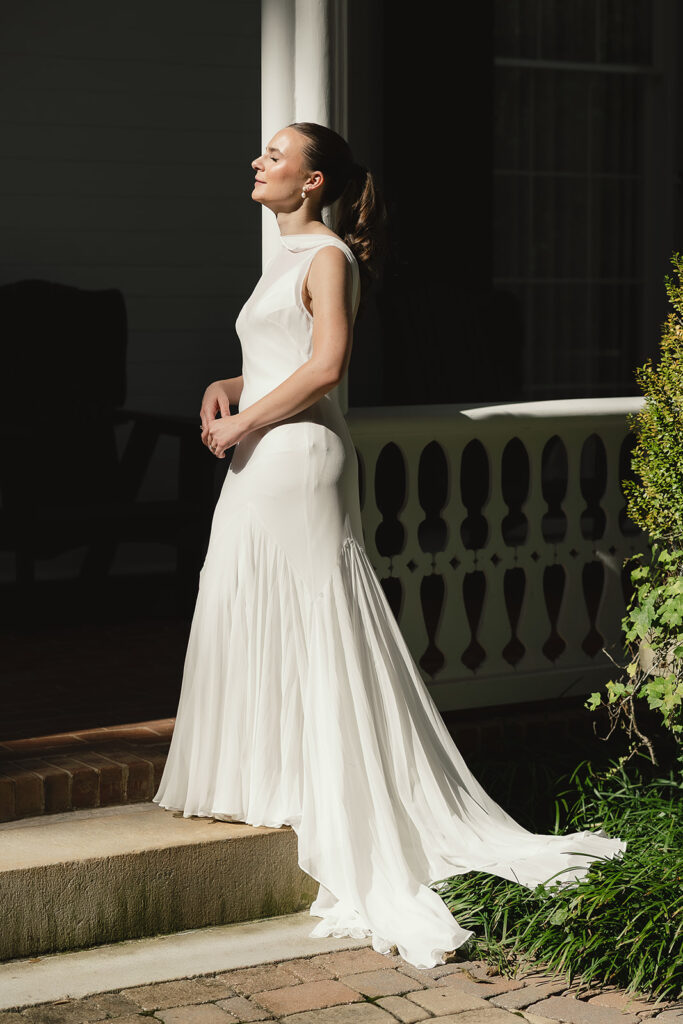 Bride standing in golden sunlight on a porch, eyes closed, savoring a quiet moment before the ceremony