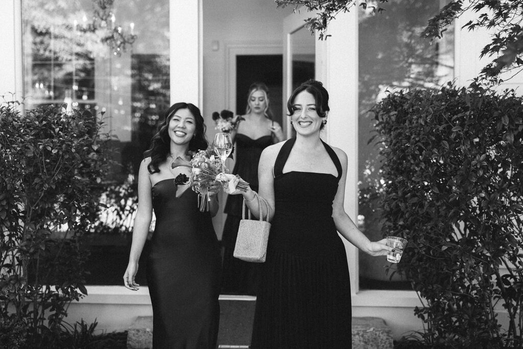 Bridesmaids in black dresses walking outside, smiling, holding flowers and drinks in a candid black and white moment