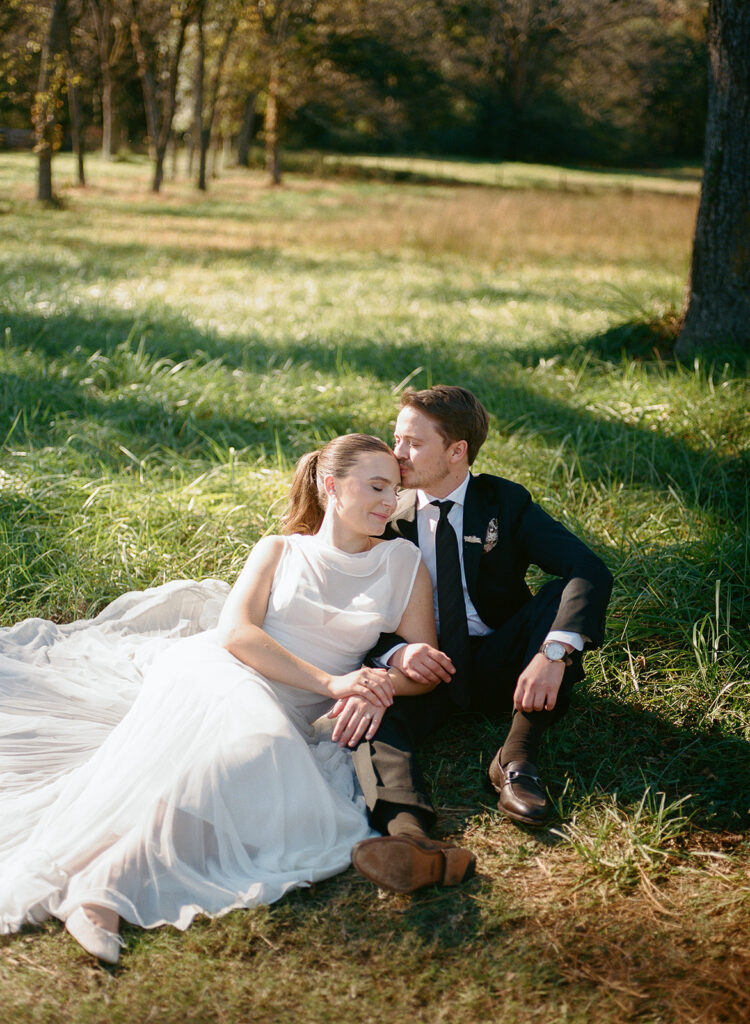 Bride and groom sitting in a sunlit field, holding hands and leaning into each other during a quiet wedding day moment