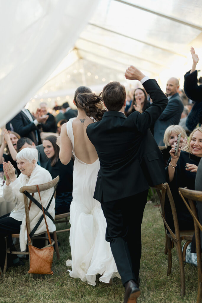 Bride and groom entering their wedding reception under a clear tent as guests cheer and clap