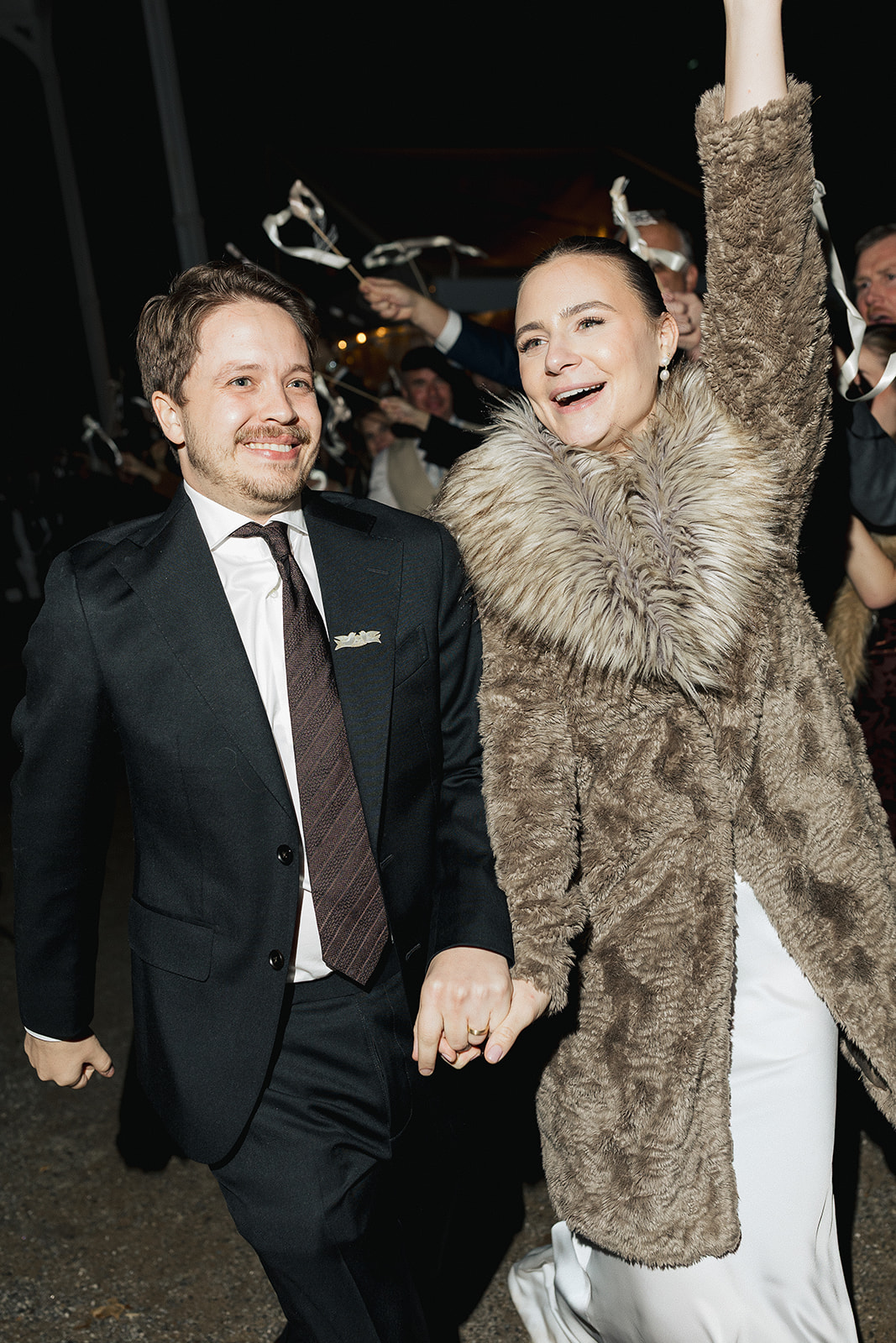 Bride and groom holding hands and smiling during their nighttime wedding exit as guests wave ribbons