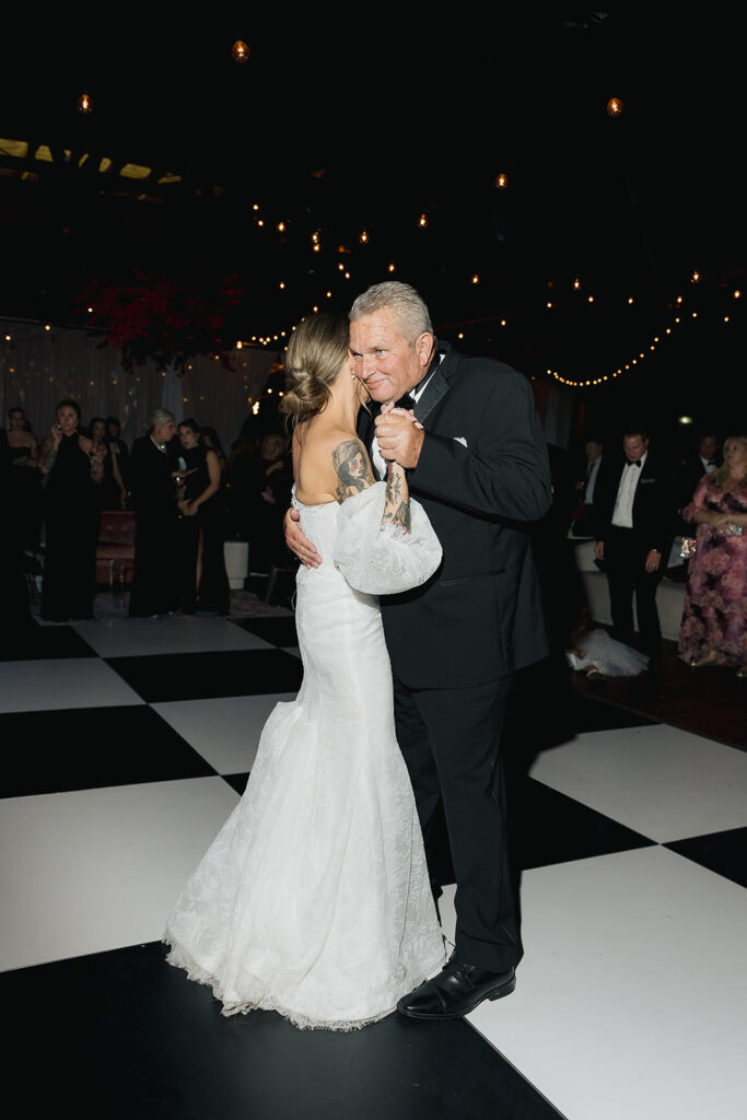 Bride dancing with an older man during the reception under warm string lights on a black and white dance floor.