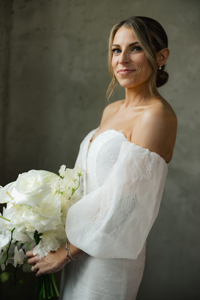 Bride holding a white bouquet, standing in soft natural light against a textured wall in an editorial wedding portrait.