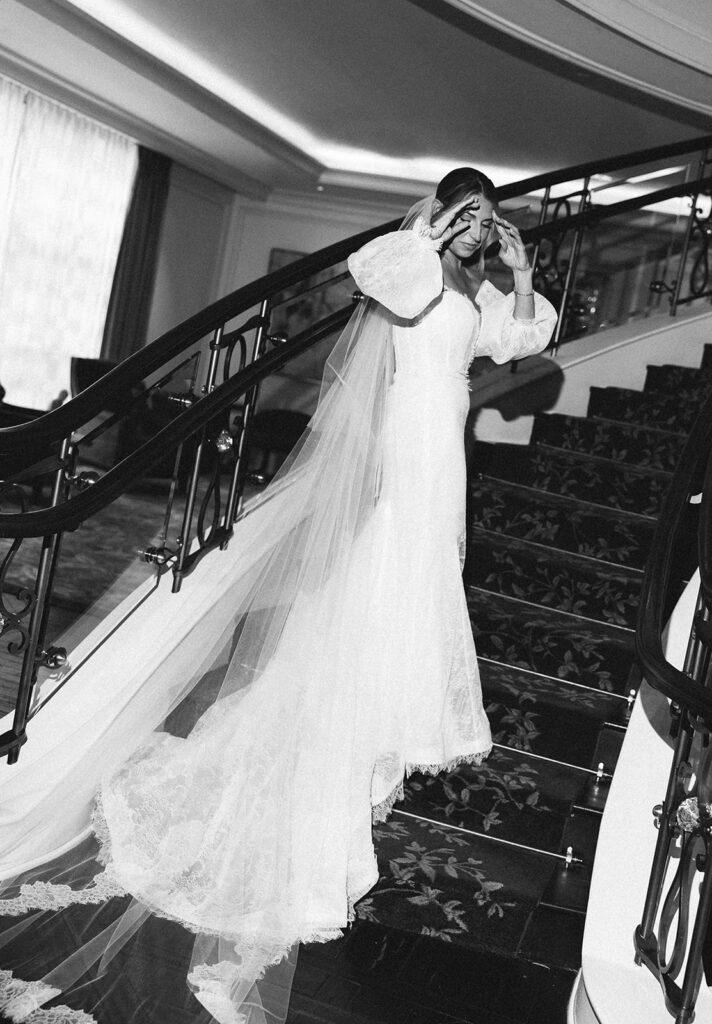 Black and white photograph of a bride standing alone on a staircase, adjusting her veil in soft window light.