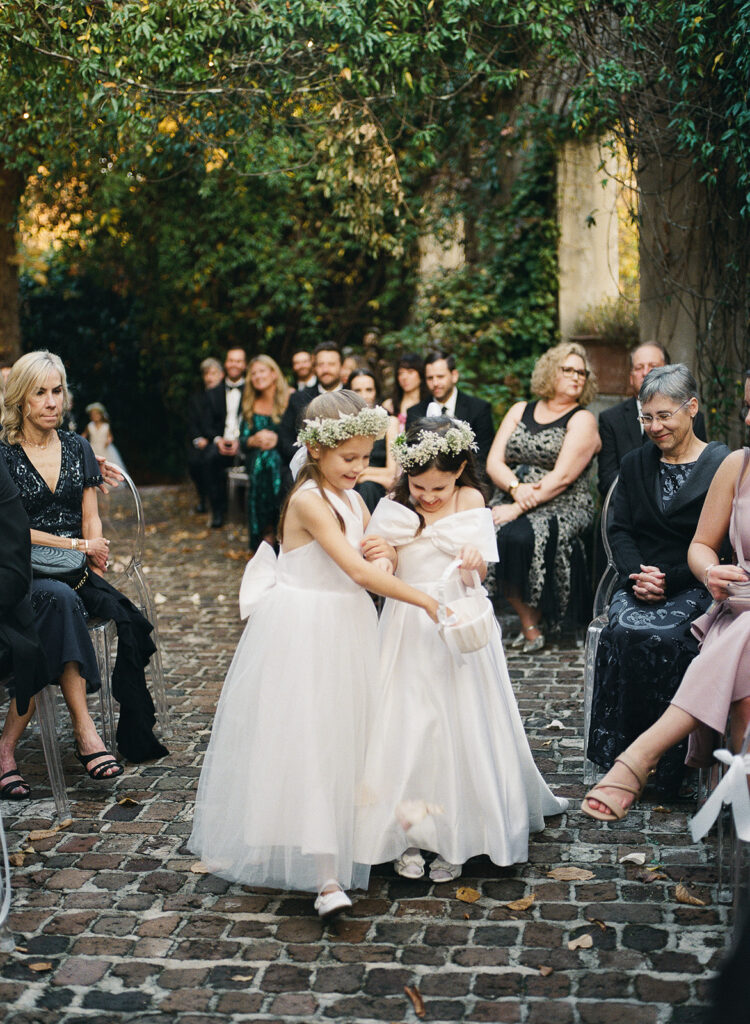 Two flower girls in white dresses walking together down a cobblestone aisle during an outdoor wedding ceremony as guests look on.