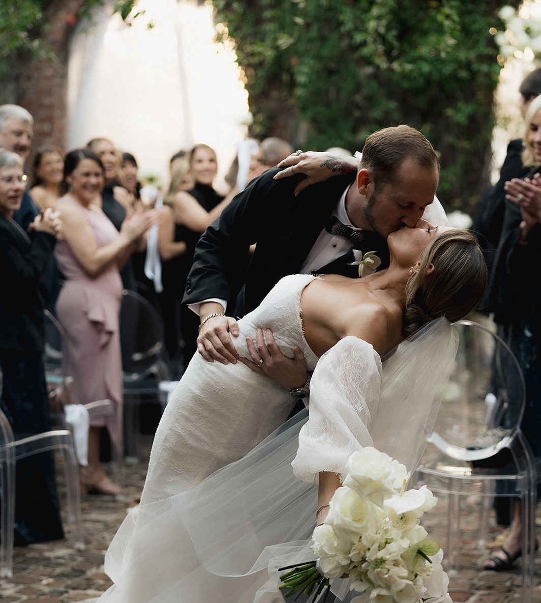 Bride and groom sharing their first kiss at the end of an outdoor garden wedding ceremony as guests applaud around them.