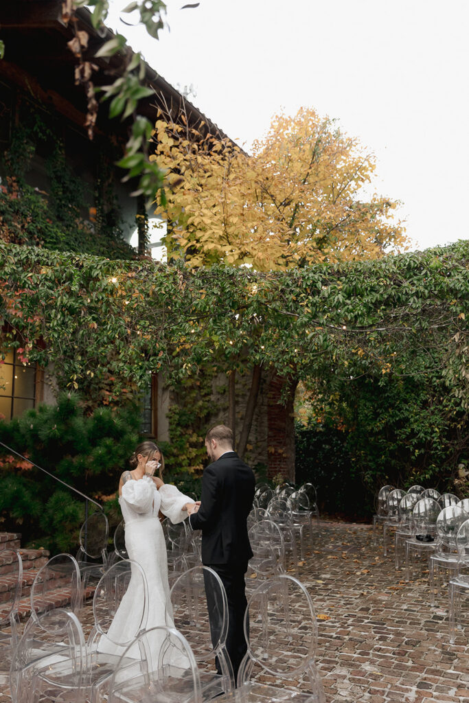 Bride and groom standing together during an outdoor ceremony as the bride wipes away tears, surrounded by empty chairs.