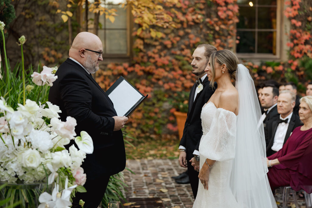 Bride and groom standing together during an outdoor wedding ceremony as the officiant reads their vows in a garden setting.