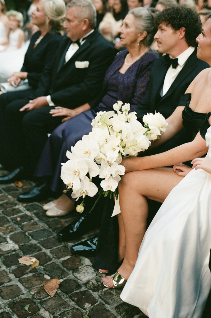 Guests seated during an outdoor wedding ceremony, holding white orchids and watching attentively as the ceremony unfolds.